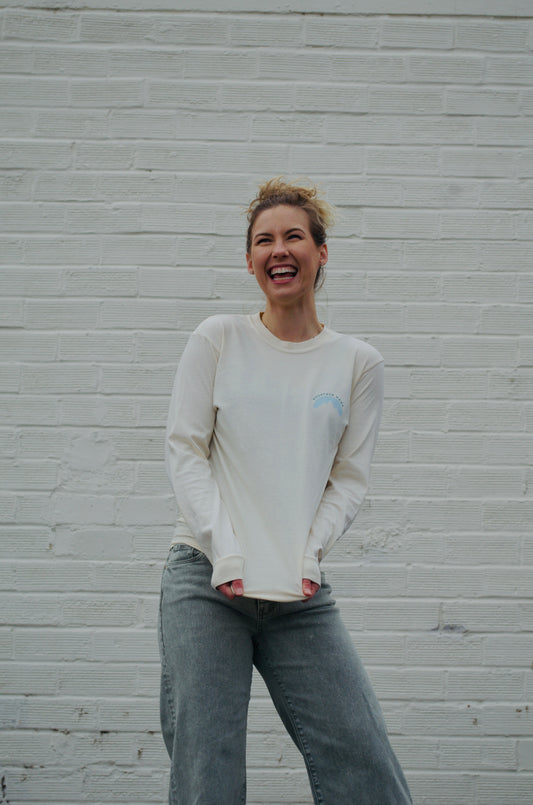 Smiling woman in a cream “Mountain Mama” long sleeve shirt with subtle chest logo, paired with faded jeans, standing in front of a white brick wall.