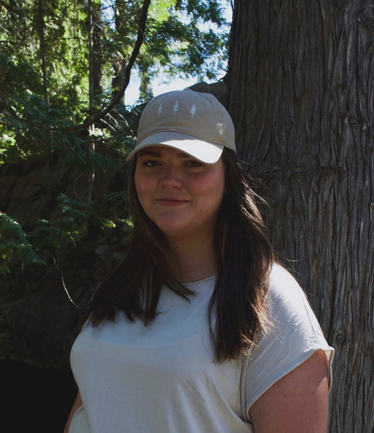 A woman wearing the Evergreen Trio Hat featuring three minimalist evergreen tree icons embroidered on the front, shown on a neutral-toned, unisex baseball cap.