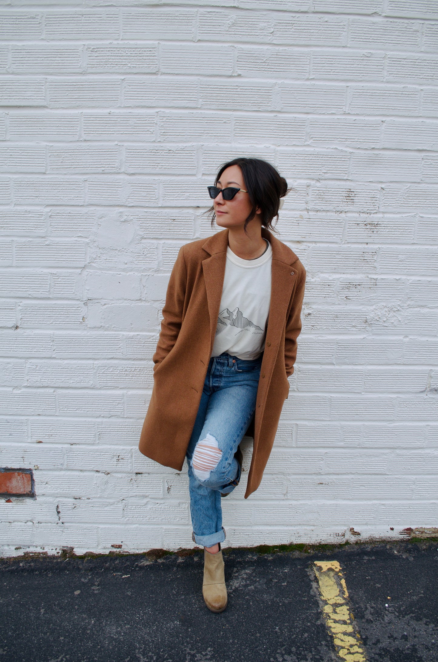 Woman wearing a camel coat over a mountain graphic T-shirt and ripped jeans, leaning casually against a white brick wall.