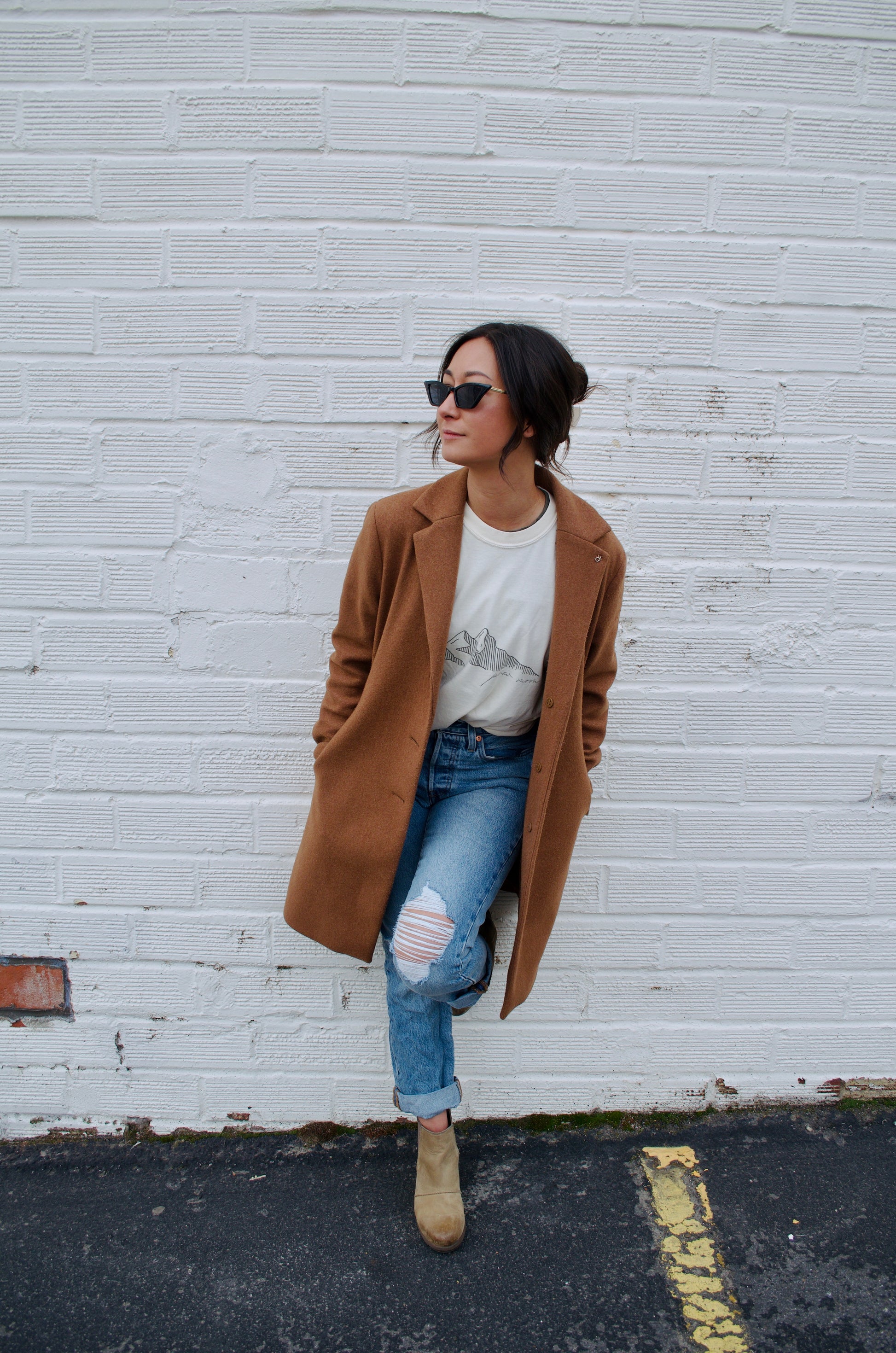 Woman wearing a camel coat over a mountain graphic T-shirt and ripped jeans, leaning casually against a white brick wall.