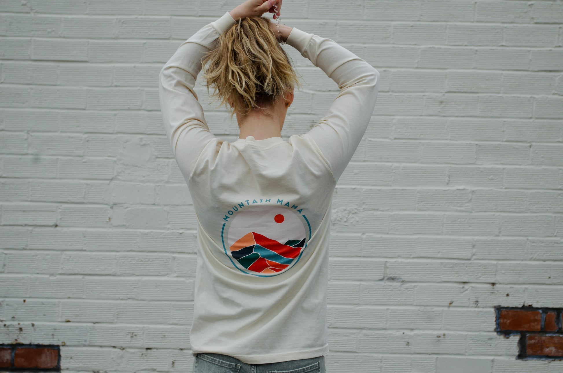 Person wearing a cream long sleeve “Mountain Mama” graphic tee, featuring a colorful mountain design on the back, standing against a white brick wall.